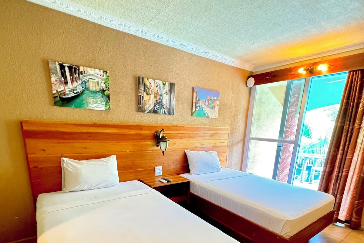 A hotel room with two beds, a wooden headboard, a nightstand, a wall-mounted lamp, and three framed pictures.