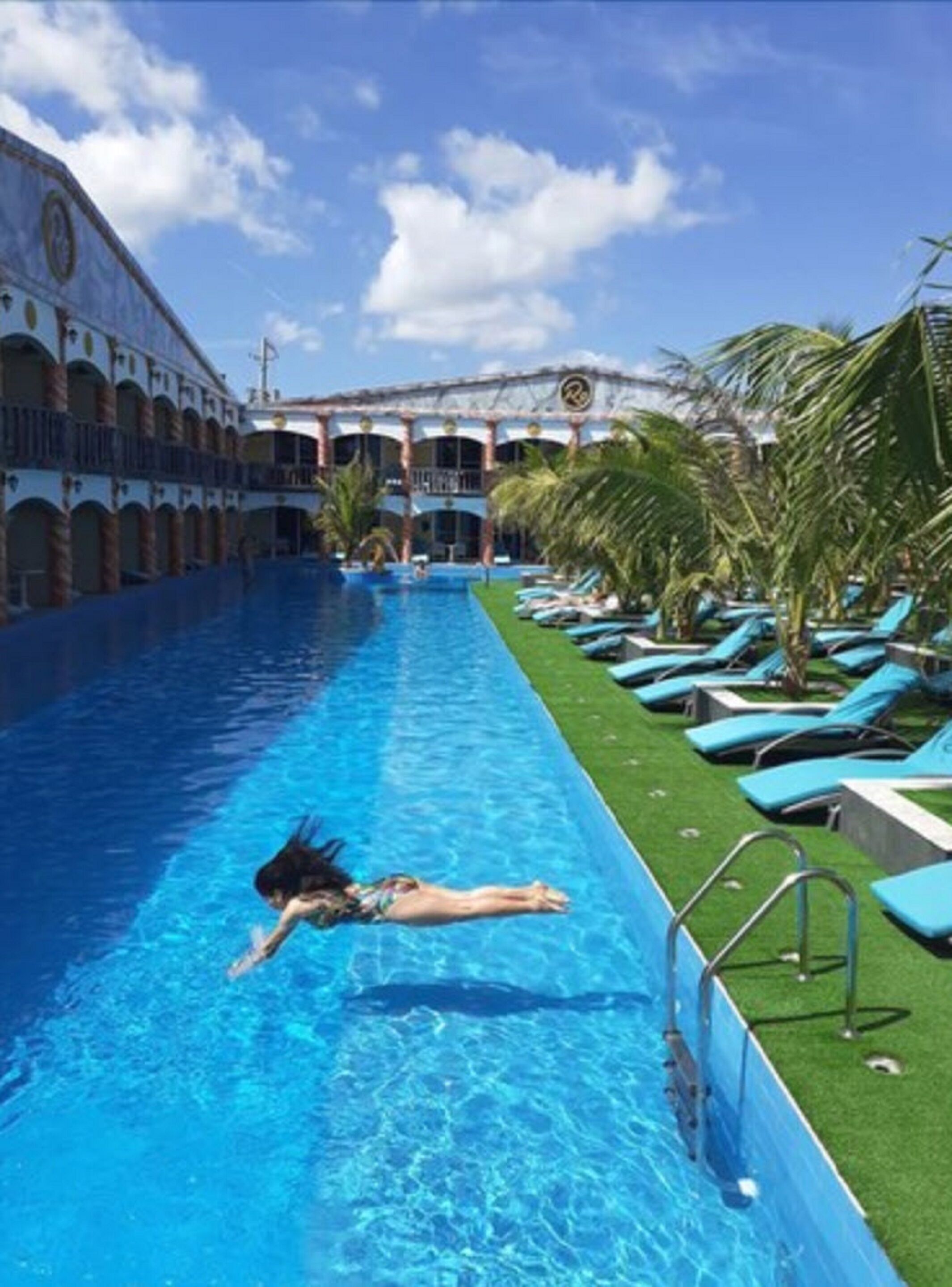 Une piscine où une personne nage, entourée de chaises longues et de palmiers.