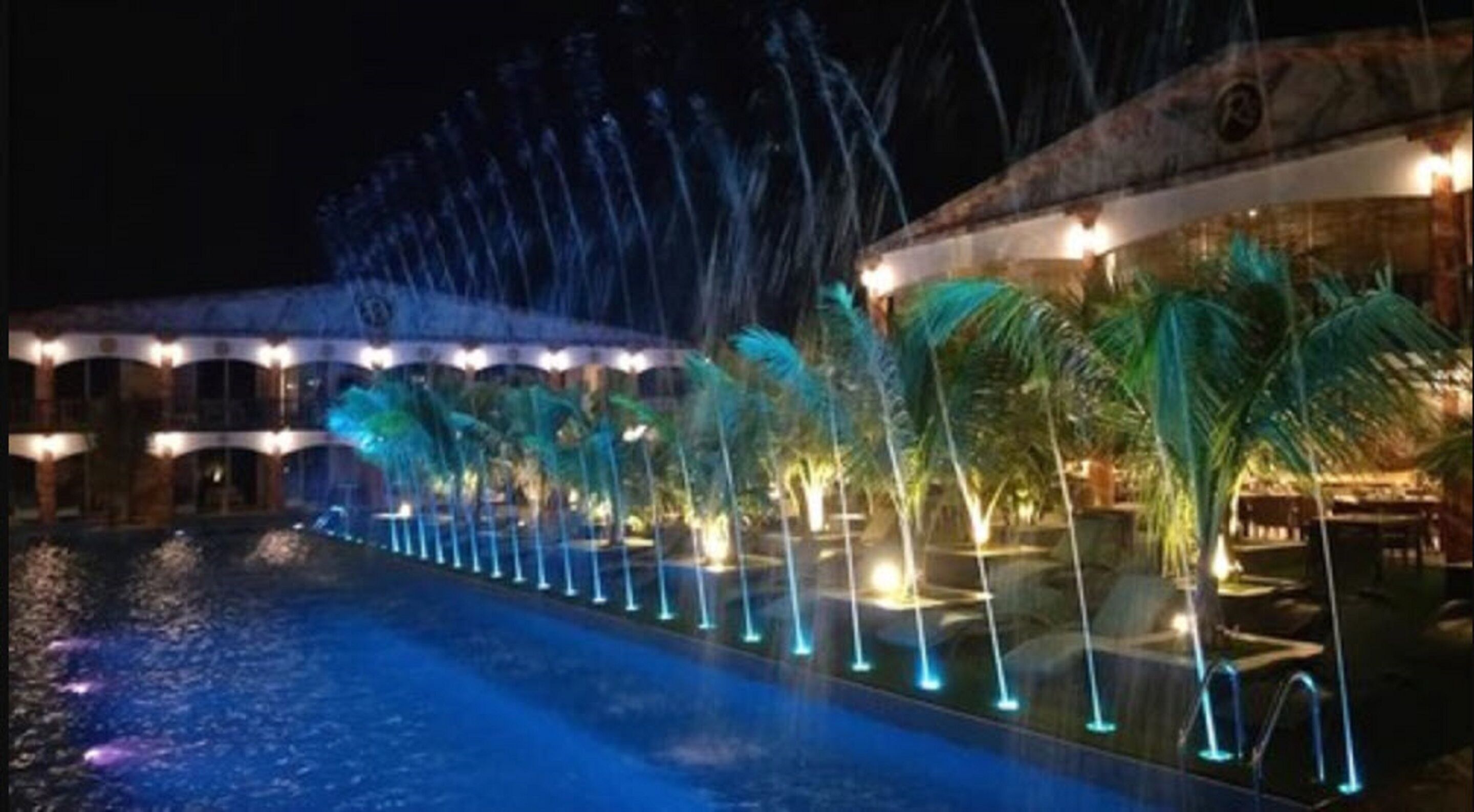 A pool with water features and palm trees, surrounded by a building with arches and lights.