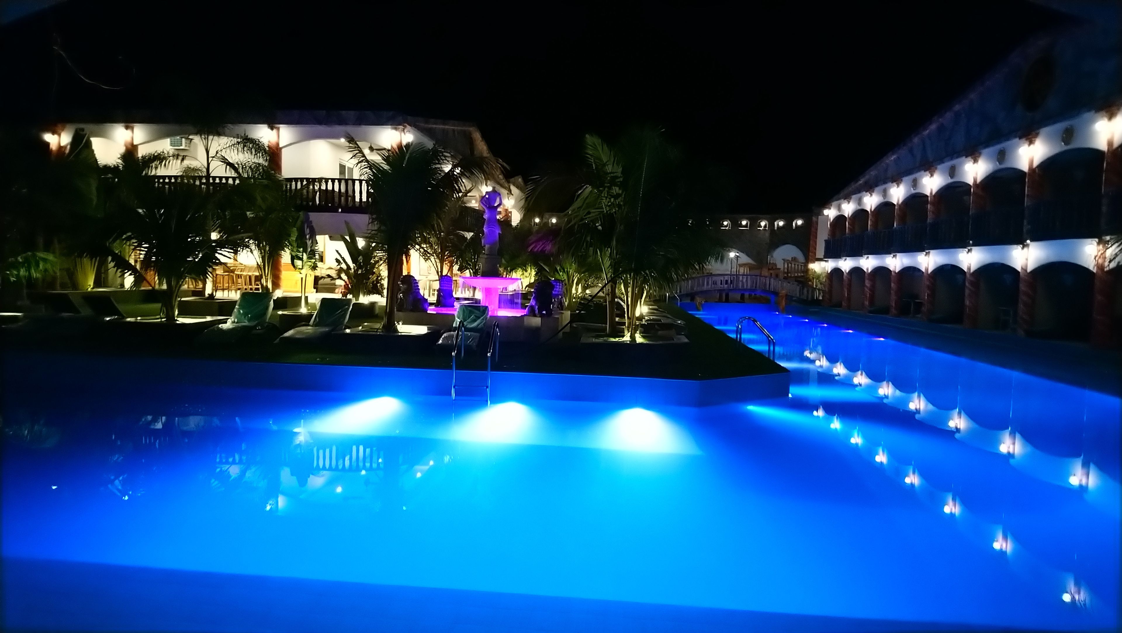 Vue nocturne d’un espace piscine d’hôtel, avec des jeux d’eau illuminés et des chaises longues.