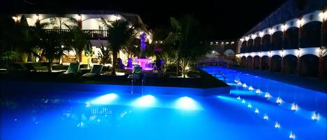 Vue nocturne d’un espace piscine d’hôtel, avec des jeux d’eau illuminés et des chaises longues.