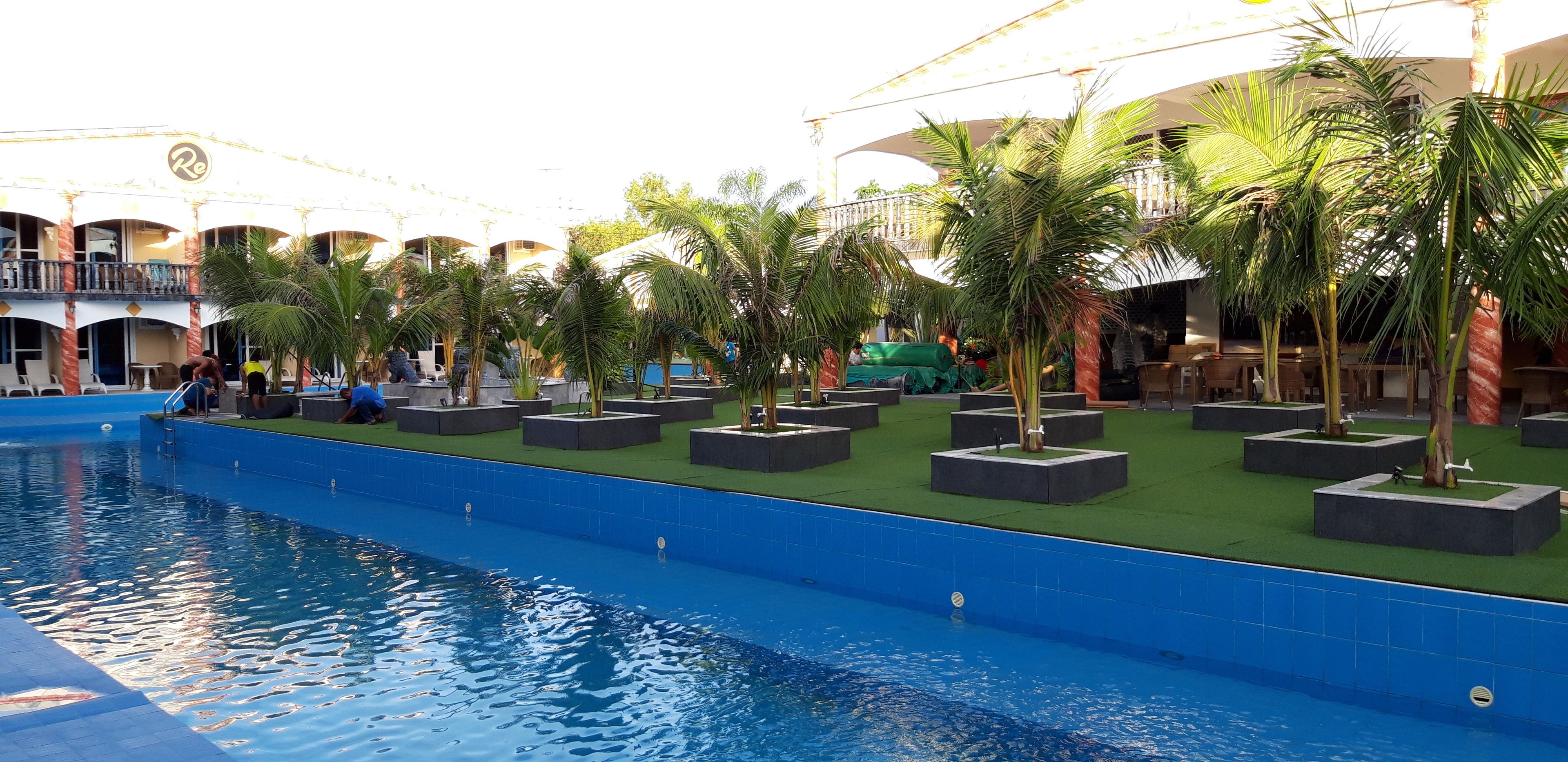 A swimming pool with a surrounding deck area, palm trees, and a building with a balcony in the background.