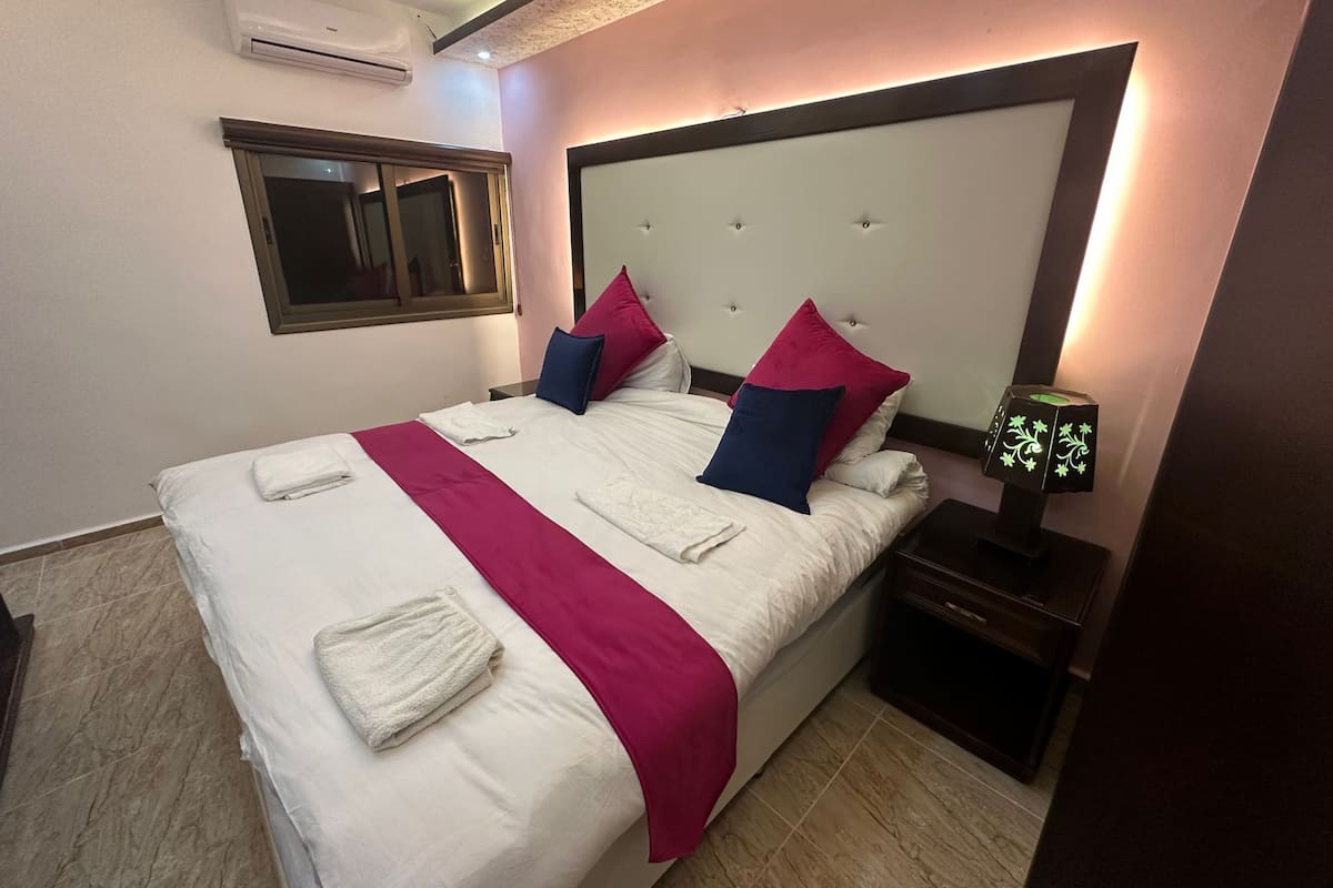 A neatly made bed with white and pink linens, a bedside table with a lamp, and a window with blinds.