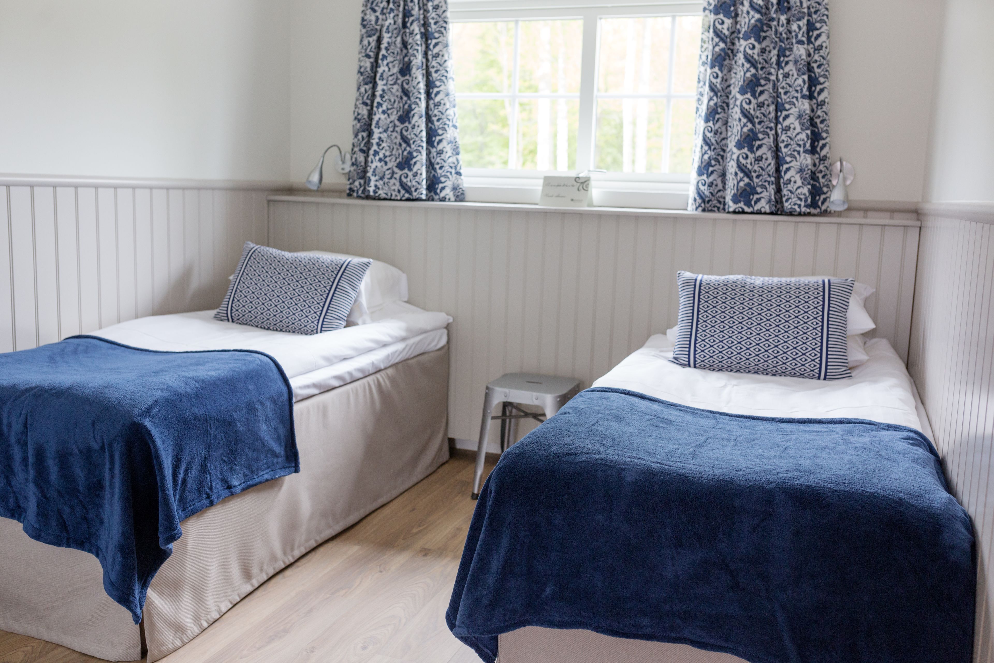 Ein Schlafzimmer mit zwei Betten, jeweils mit blauer Decke und passenden Kissen. Es gibt ein Fenster mit gemusterten Vorhängen, einen kleinen Nachttisch und eine Heizung über den Betten.