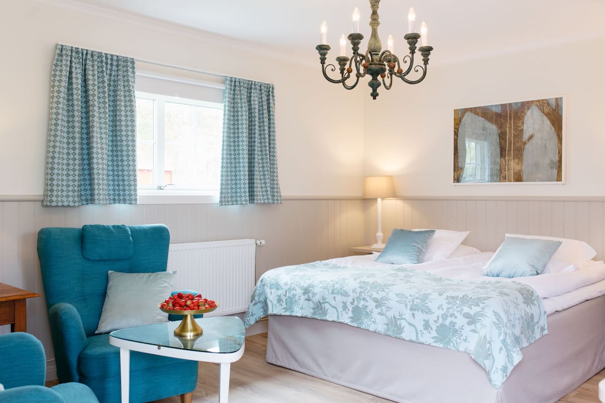 Ein Schlafzimmer mit einem Bett, einem Sessel, einem Tisch mit einer Vase, einer Chandelierleuchtre und einem Fenster mit Vorhängen.