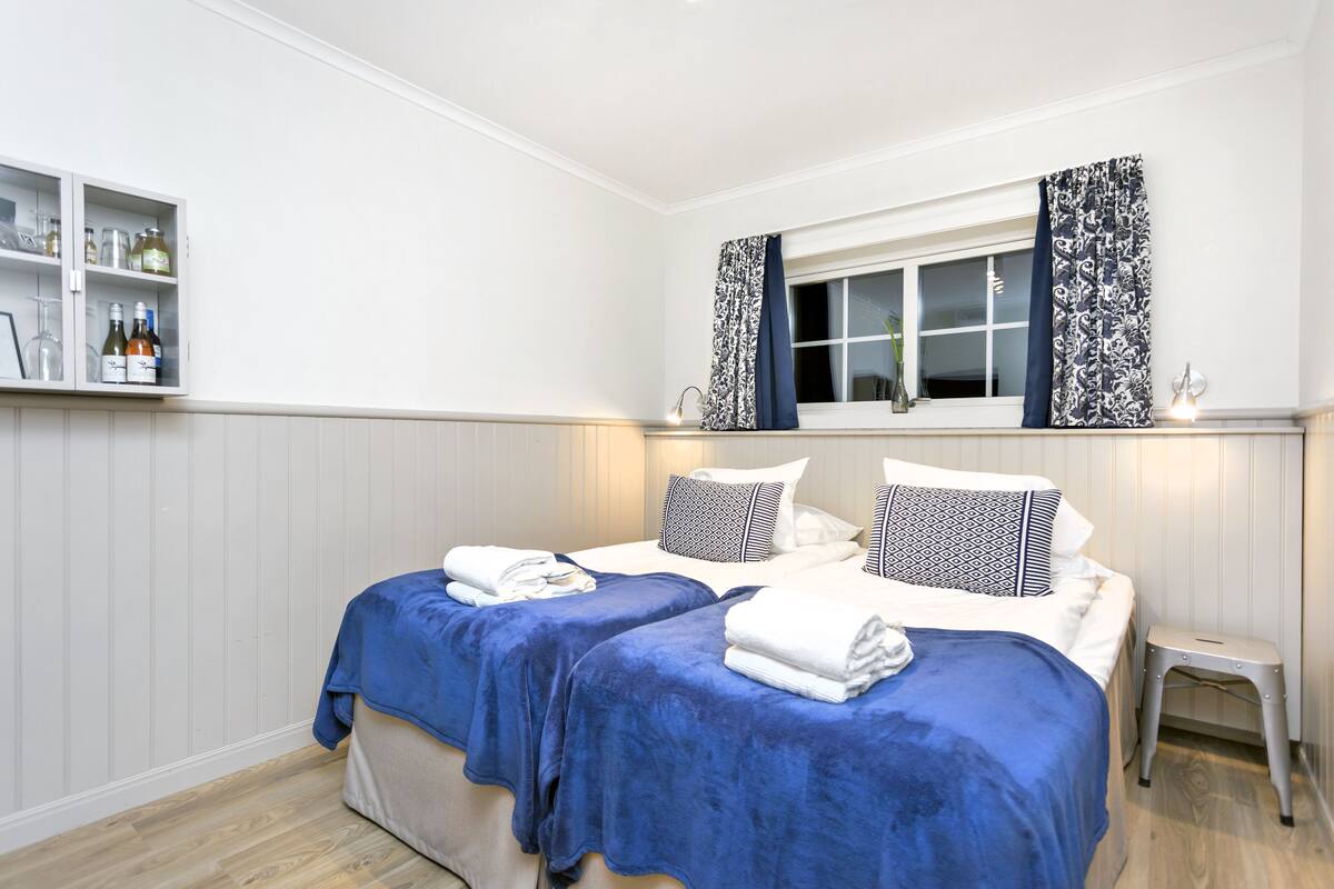 Ein Hotelzimmer mit zwei Betten, einem kleinen Nachttisch, einem Fenster mit gemusterten Vorhängen und einem Regal mit Flaschen.