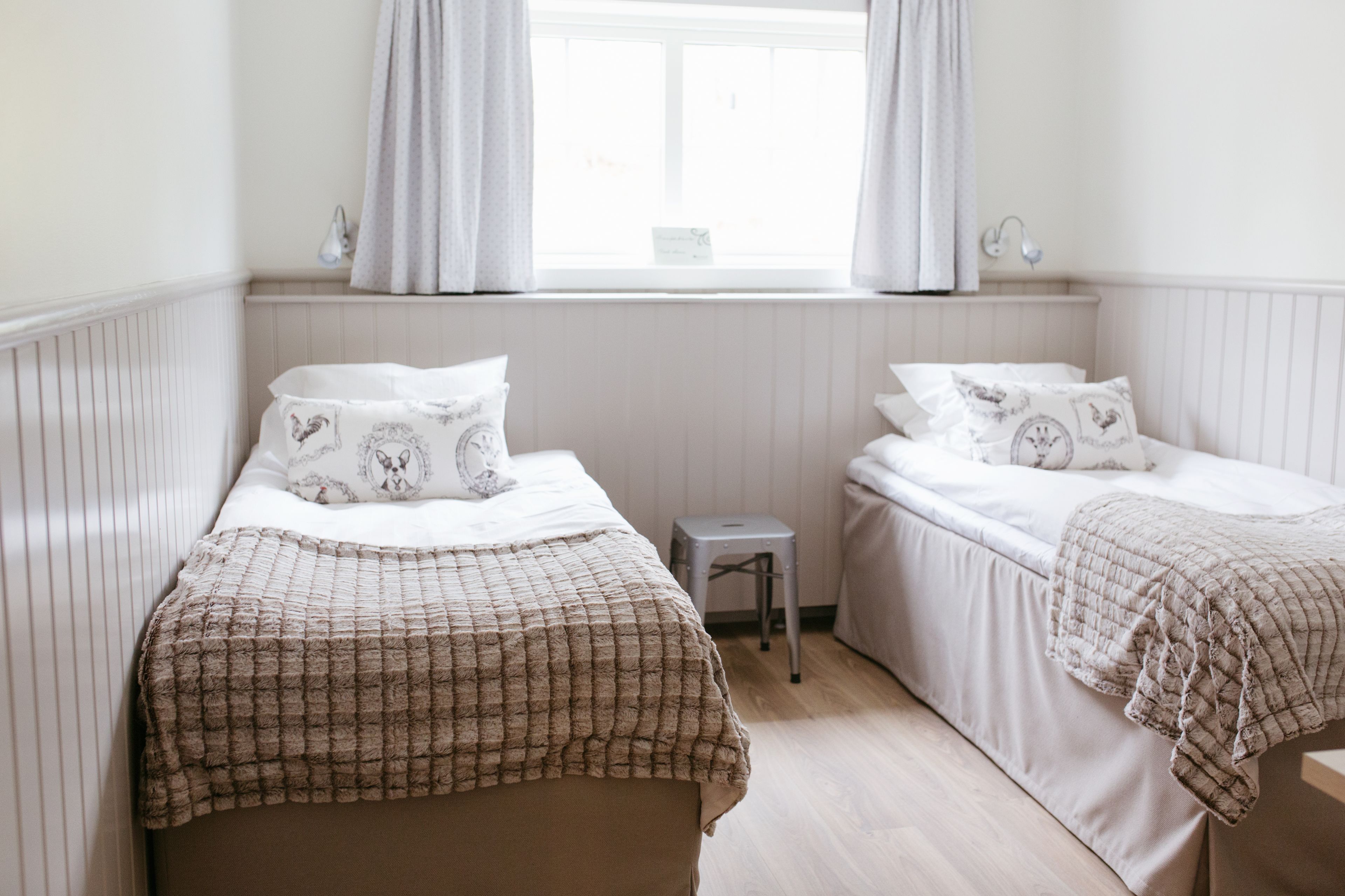 Habitación doble con dos camas, una ventana con cortinas, un taburete pequeño y un radiador.