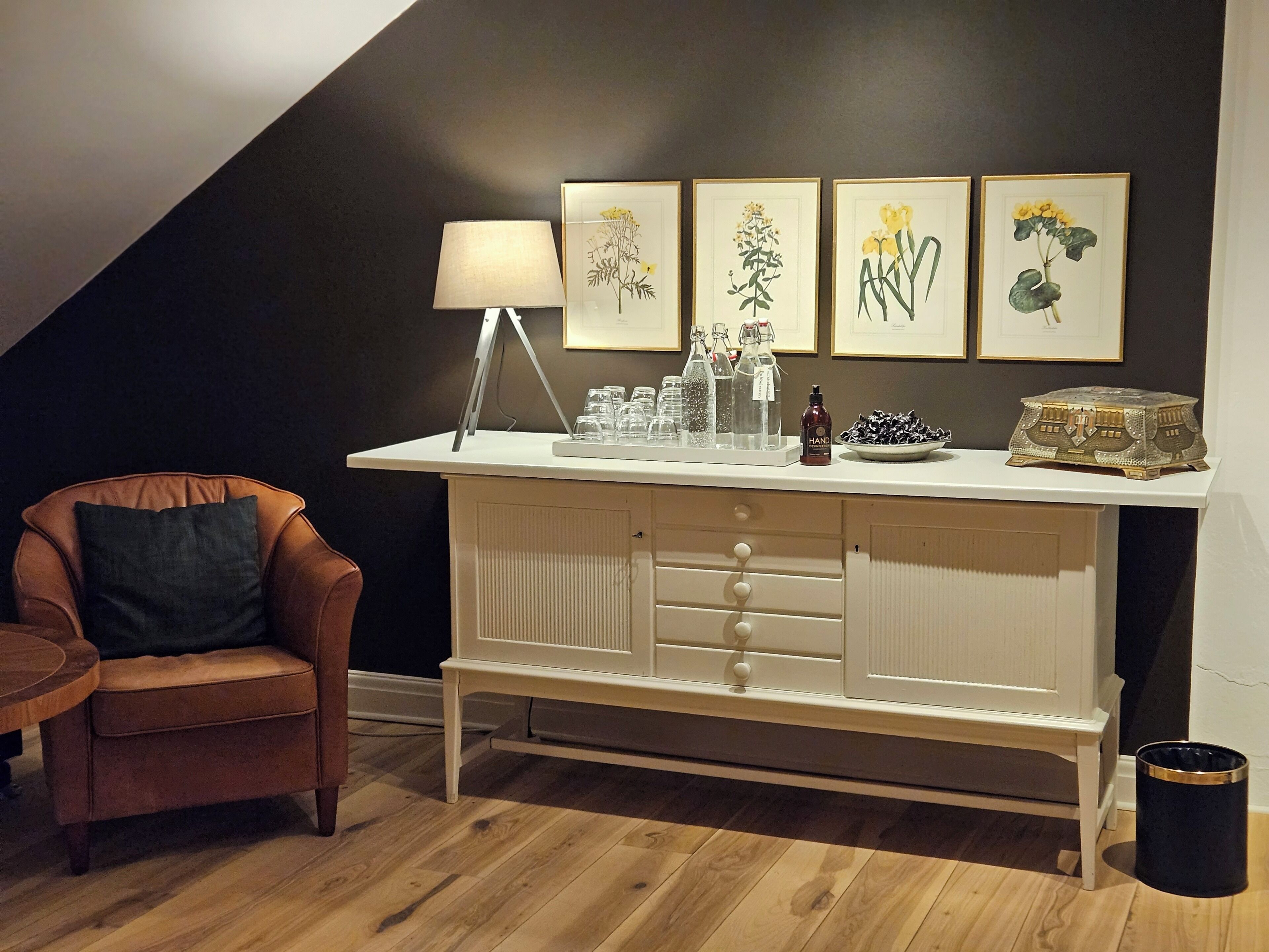 Ett rum med ett vitt skåp, en brun läderfåtölj och en lampa. På väggen hänger inramade botaniska illustrationer.