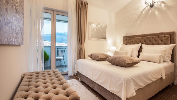 A bedroom with a large bed, a tufted bench, a view of the outdoors, and a chandelier. - Apartments Toni Relax (Split)