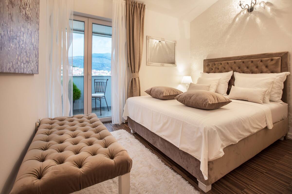 A bedroom with a large bed, a tufted bench, a view of the outdoors, and a chandelier.