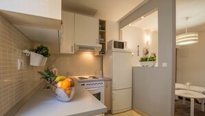 A compact kitchen with a microwave, stove, and refrigerator. A fruit bowl is on the counter, and there is a dining area with a table and chairs. - Apartments Toni Relax (Split)