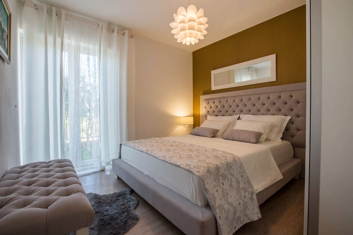 A bedroom with a large bed, a tufted headboard, a bedside lamp, and a window with sheer curtains.