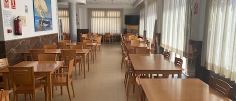 A spacious dining area with multiple wooden tables and chairs, a tiled floor, and large windows with curtains.