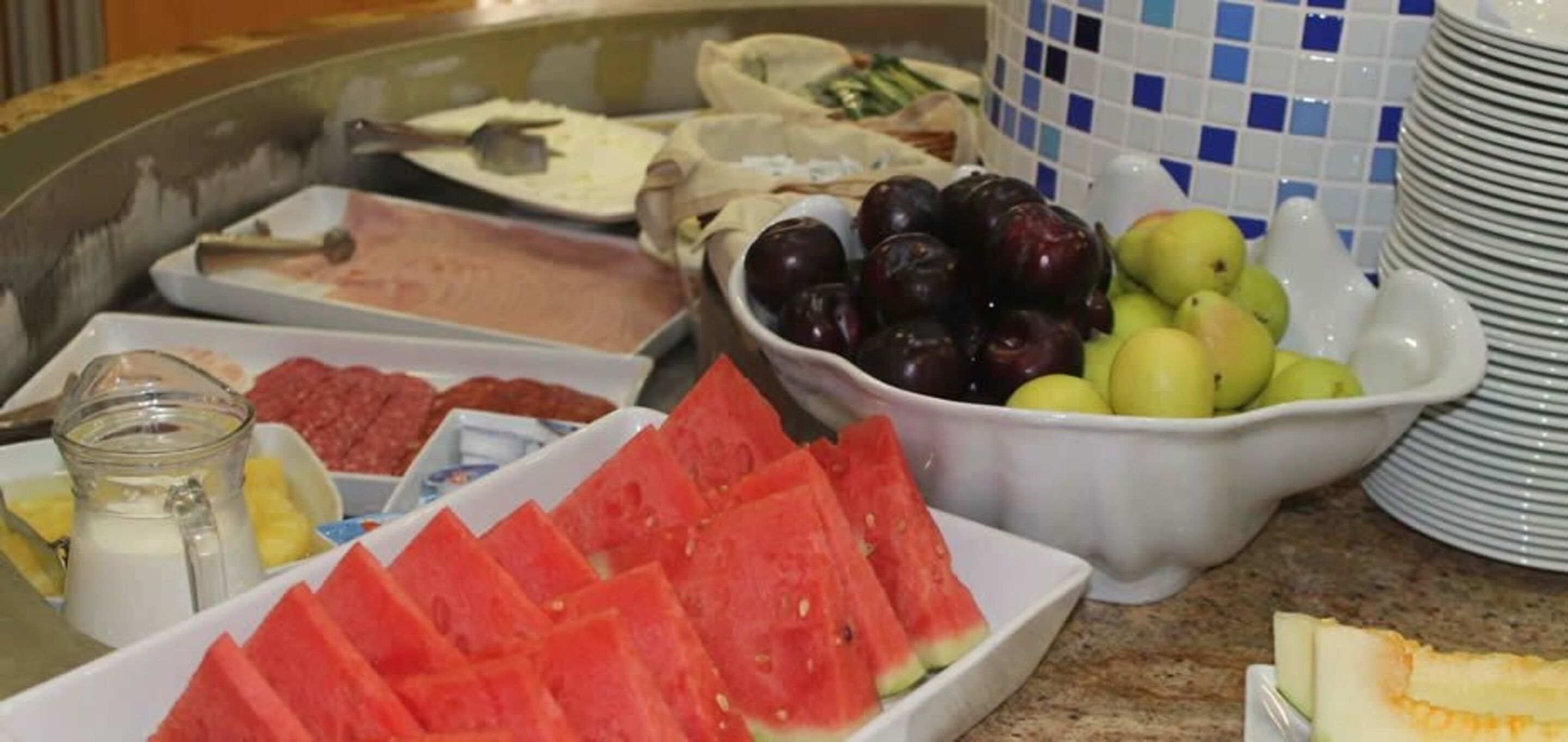 A buffet with various food items including watermelon slices, plums, apples, and a bowl of yogurt.