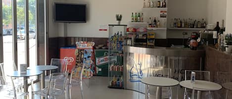 Un hall de hotel con mesas y sillas de vidrio transparente, una zona de bar con varias botellas, y un televisor montado en la pared.