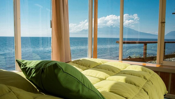 A bed with green and yellow bedding, a window with a view of the sea and a mountain, and a wooden railing outside.