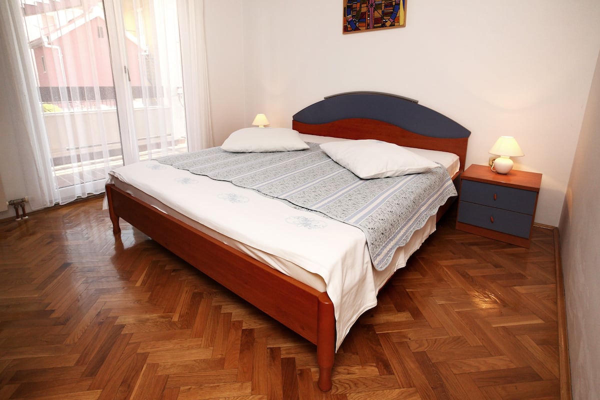 Quarto com cama de madeira, duas mesas de cabeceira com abajures, uma janela com cortinas leves e um pequeno objeto decorativo na parede.