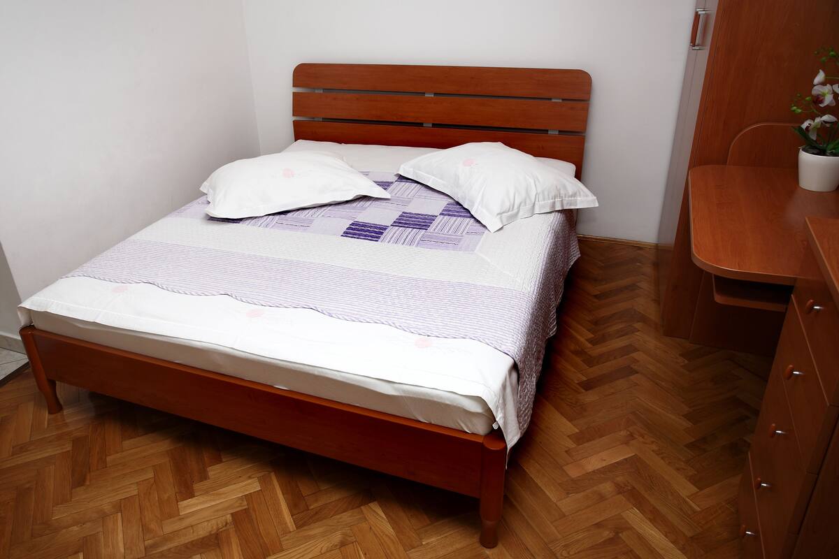Uma cama bem arrumada com cabeceira de madeira, uma cômoda de madeira à direita e um piso de madeira em padrão herringbone.