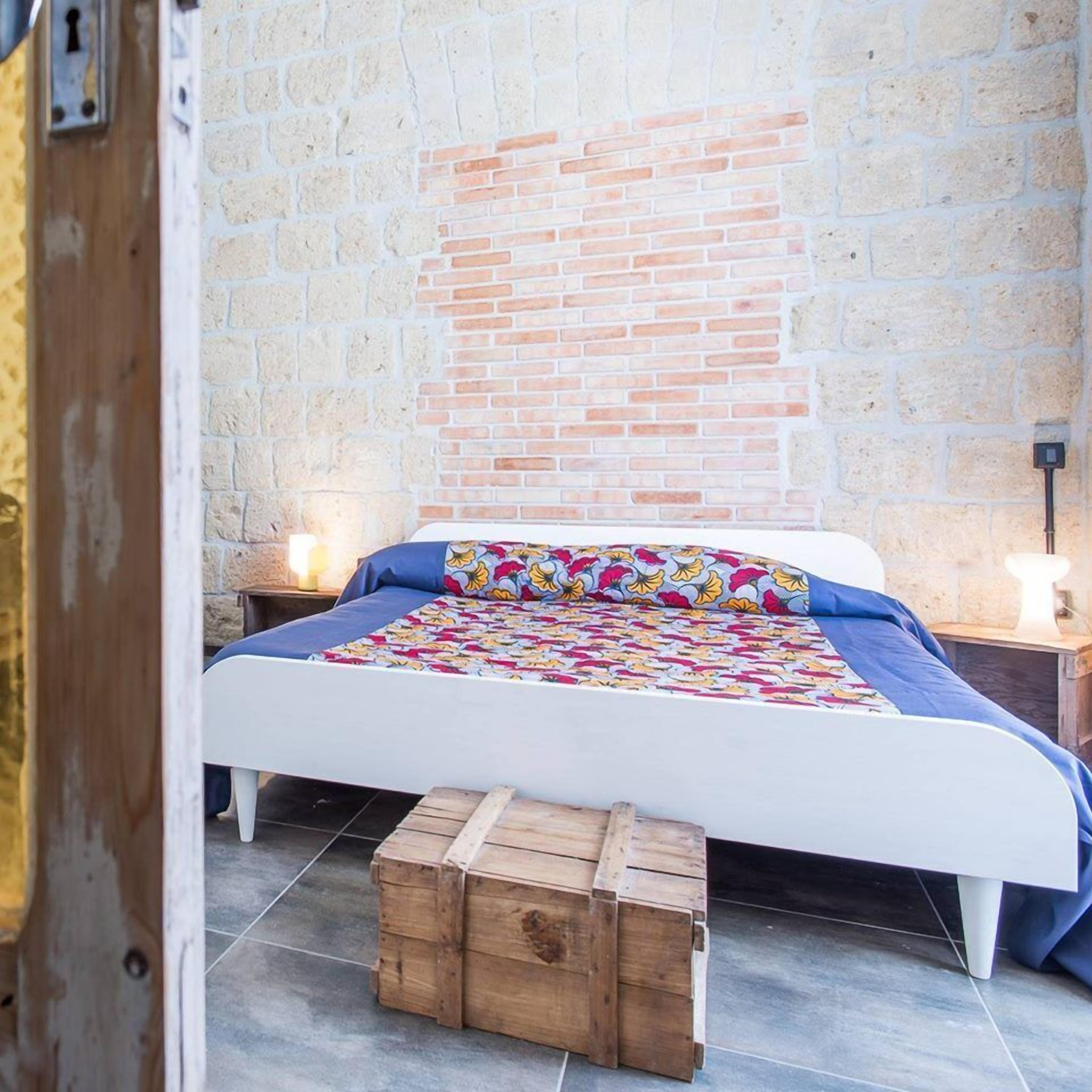 A bedroom with a brick wall, a bed with a colorful floral bedspread, two bedside tables with lamps, and a wooden chest of drawers.