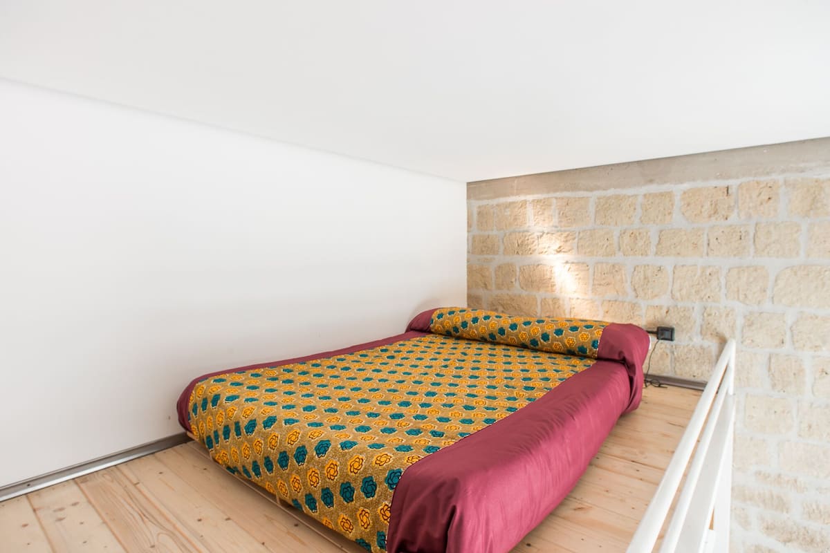 A small, single-bed room with a wooden floor, a patterned bedspread, and a stone wall.