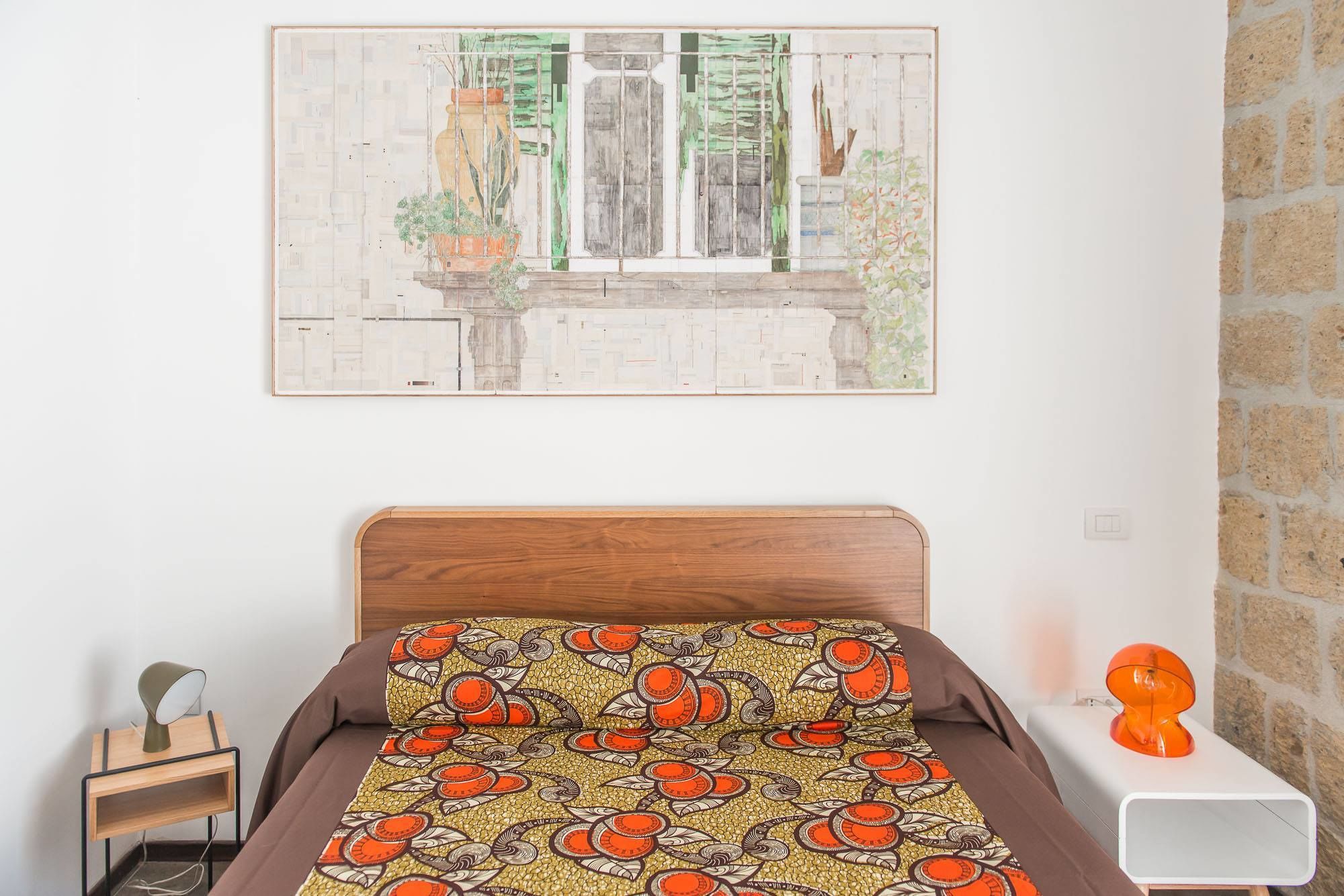 A bedroom with a bed, a nightstand, a lamp, a decorative orange object, and a painting of a window scene.