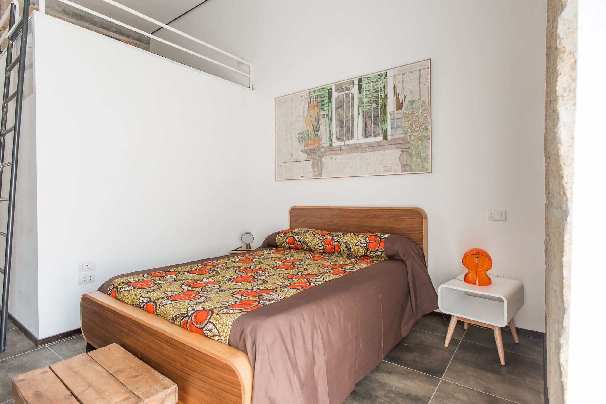 A bedroom with a wooden bed, a small bedside table with a lamp, a window with a view of buildings, and a wall-mounted painting.