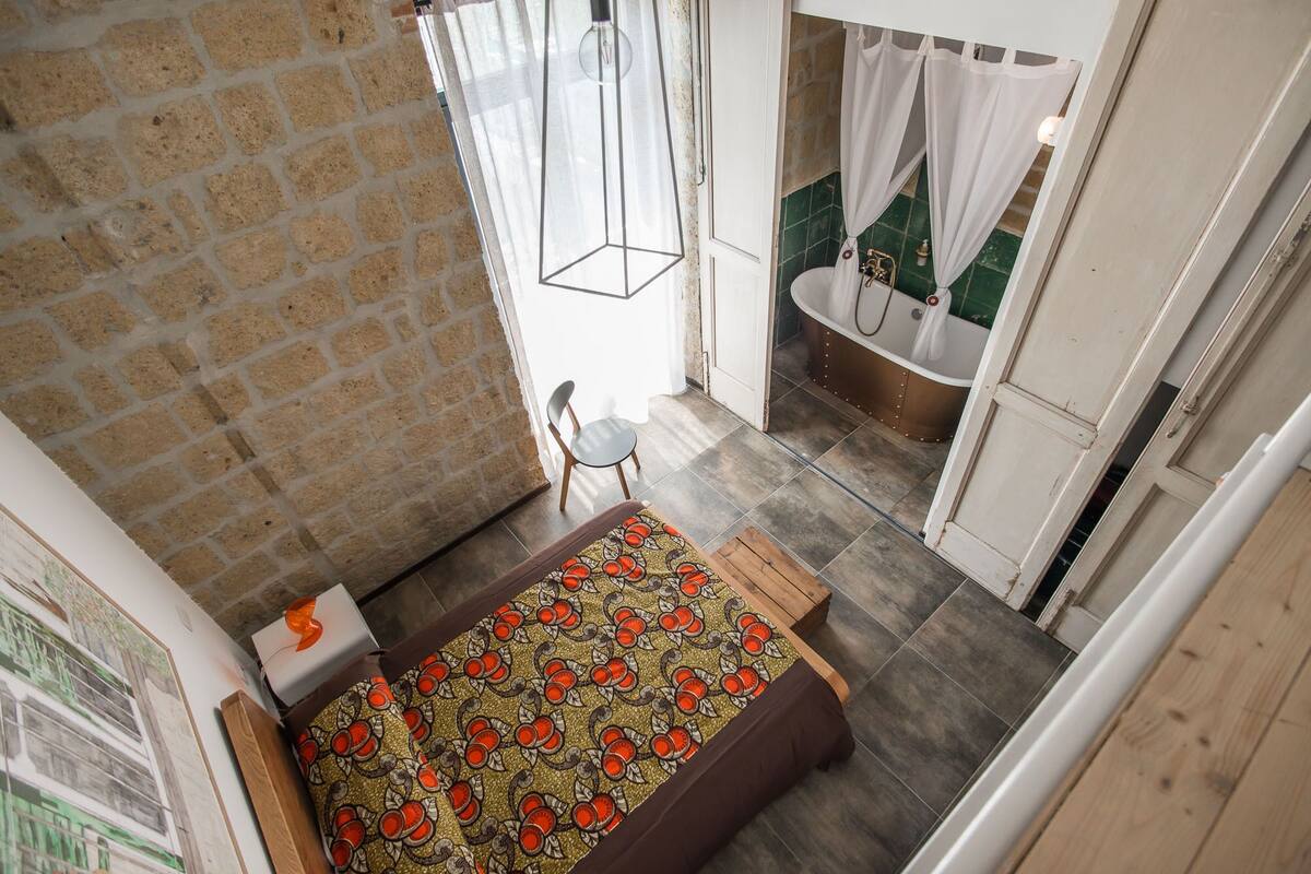 A bedroom with a bed, a chair, a window, and a bathroom with a bathtub.