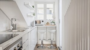 A modern kitchen with white cabinetry, a built-in oven, a microwave, and a sink. There are two white bar stools and a window with a view of the city. - Romantic Gem in Alcântara (Lisboa)