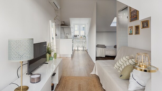 A modern, well-lit living room with a TV, sofa, and decorative items. A kitchen with a dining area is visible in the background. - Romantic Gem in Alcântara (Lisboa)