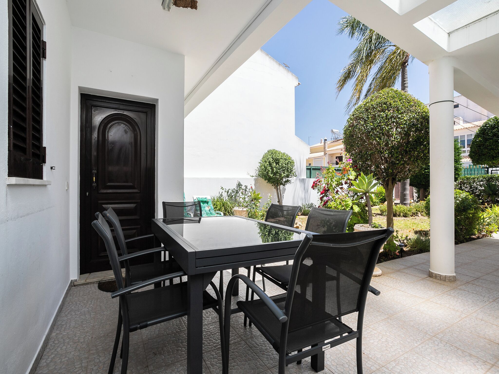 Eine Terrasse mit Tisch und Stühlen, einer Tür und Blick auf einen Garten mit verschiedenen Pflanzen und einer Palme.