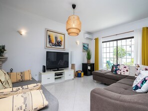 A living room with a flat-screen TV, a sofa, a coffee table, and a large window with yellow curtains.
