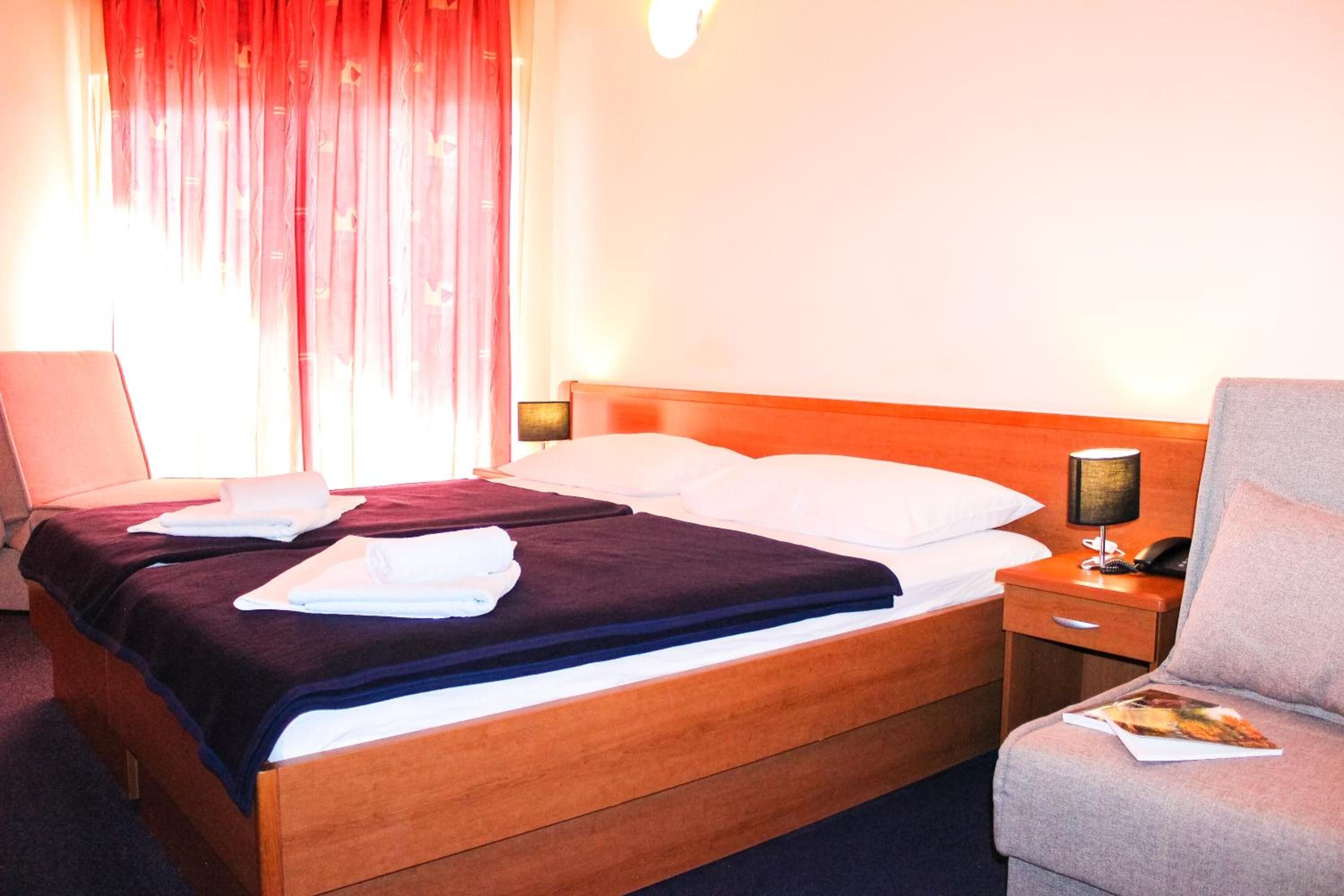 Quarto de hotel com cama de madeira, almofadas brancas, mesa de cabeceira com abajur e uma cadeira.