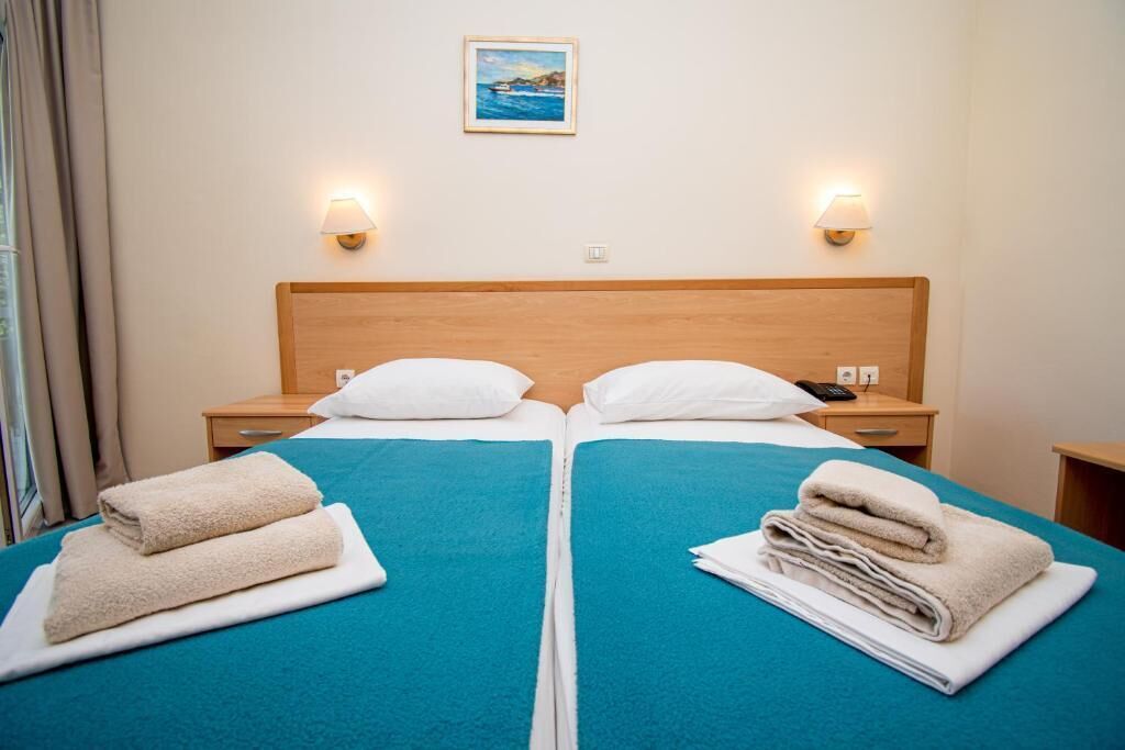 Habitación de hotel con dos camas, cada una con cubrecama azul y almohadas blancas. Hay dos lámparas de noche, un cuadro en la pared y una ventana con cortinas.