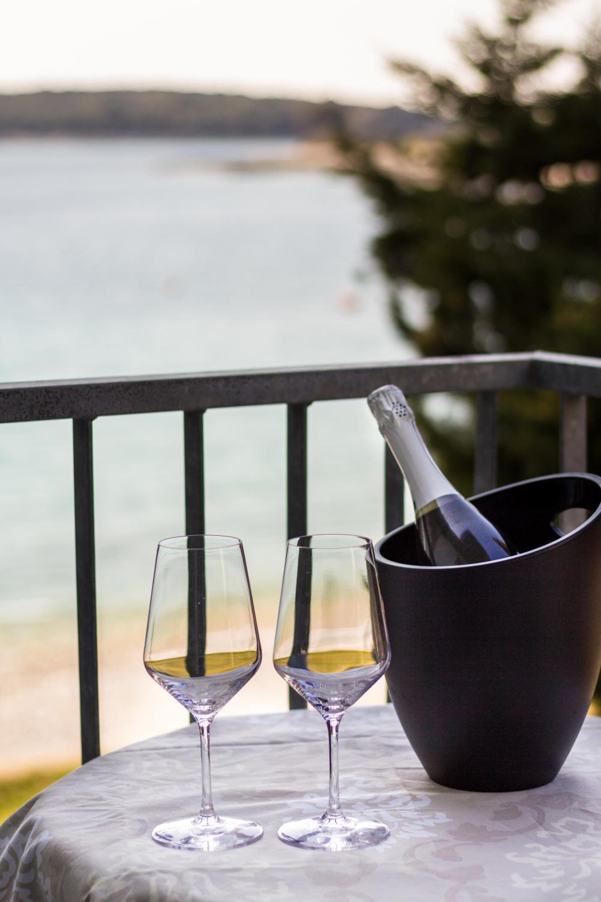 A table with two wine glasses and a champagne bottle on a balcony overlooking a body of water.