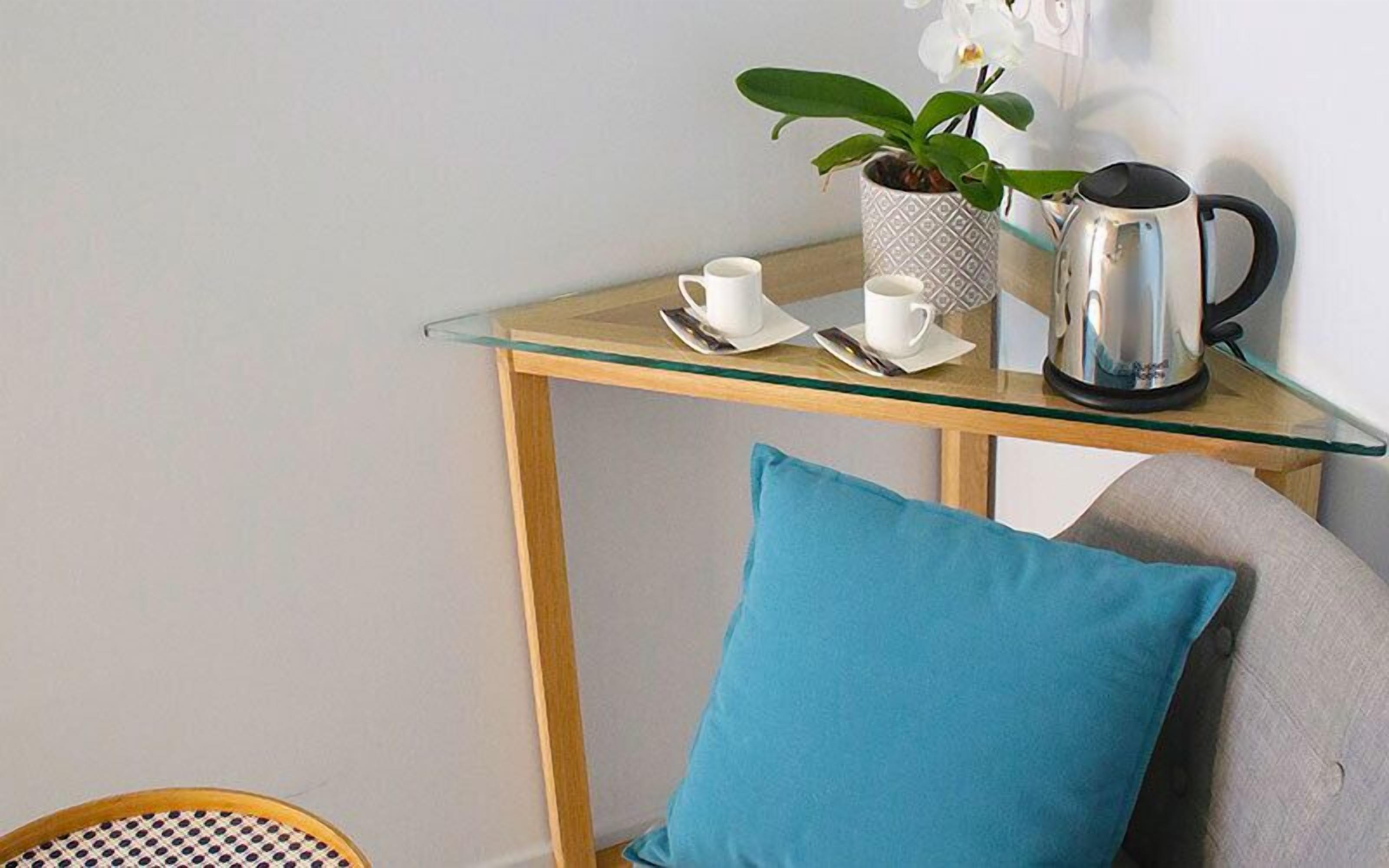 Una pequeña mesa de vidrio con una taza blanca, un platillo y una tetera eléctrica plateada.