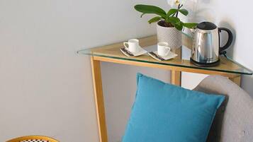 Una pequeña mesa de vidrio con una taza blanca, un platillo y una tetera eléctrica plateada.