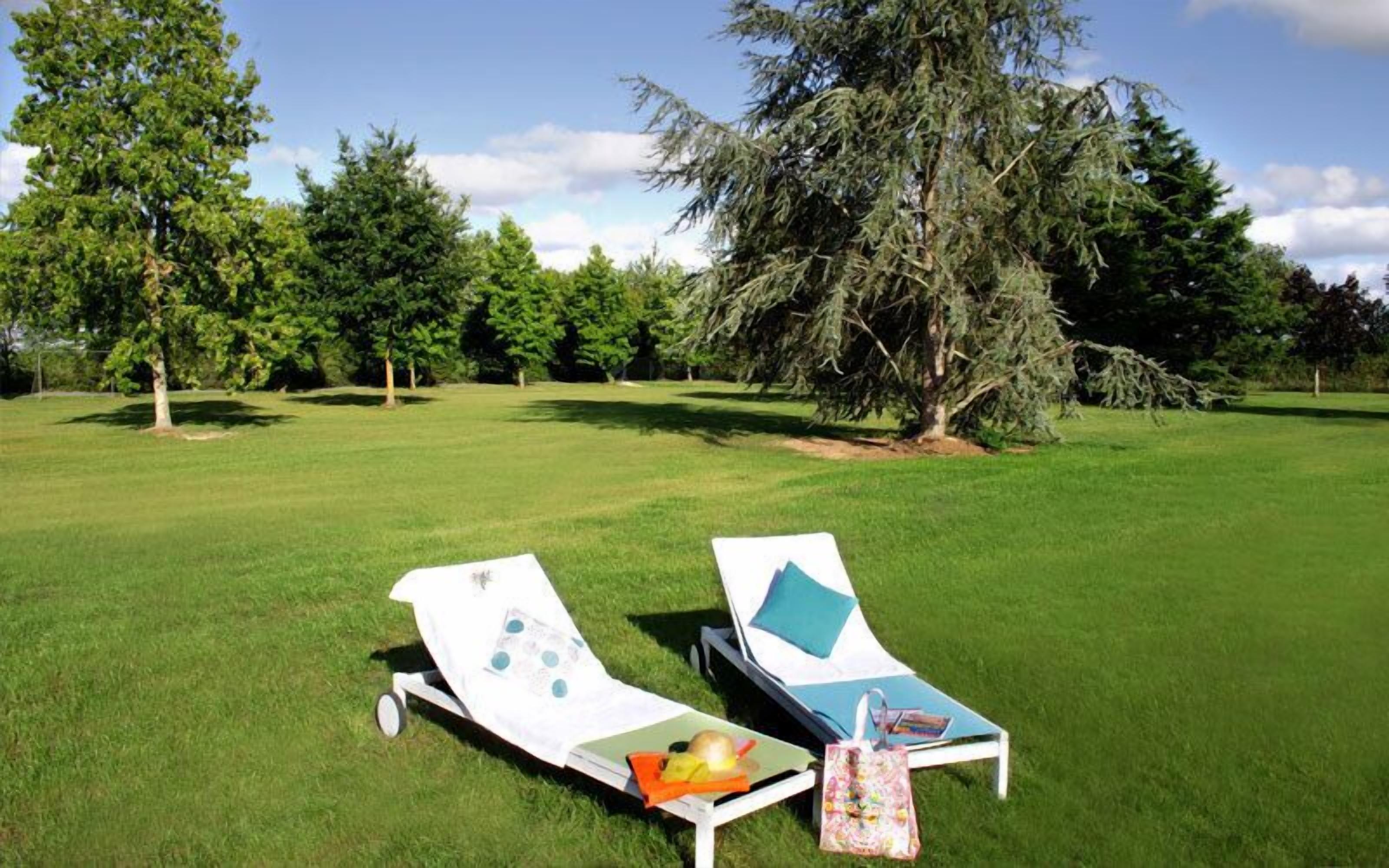 Two sun loungers with towels and a bag on the grass, surrounded by trees.