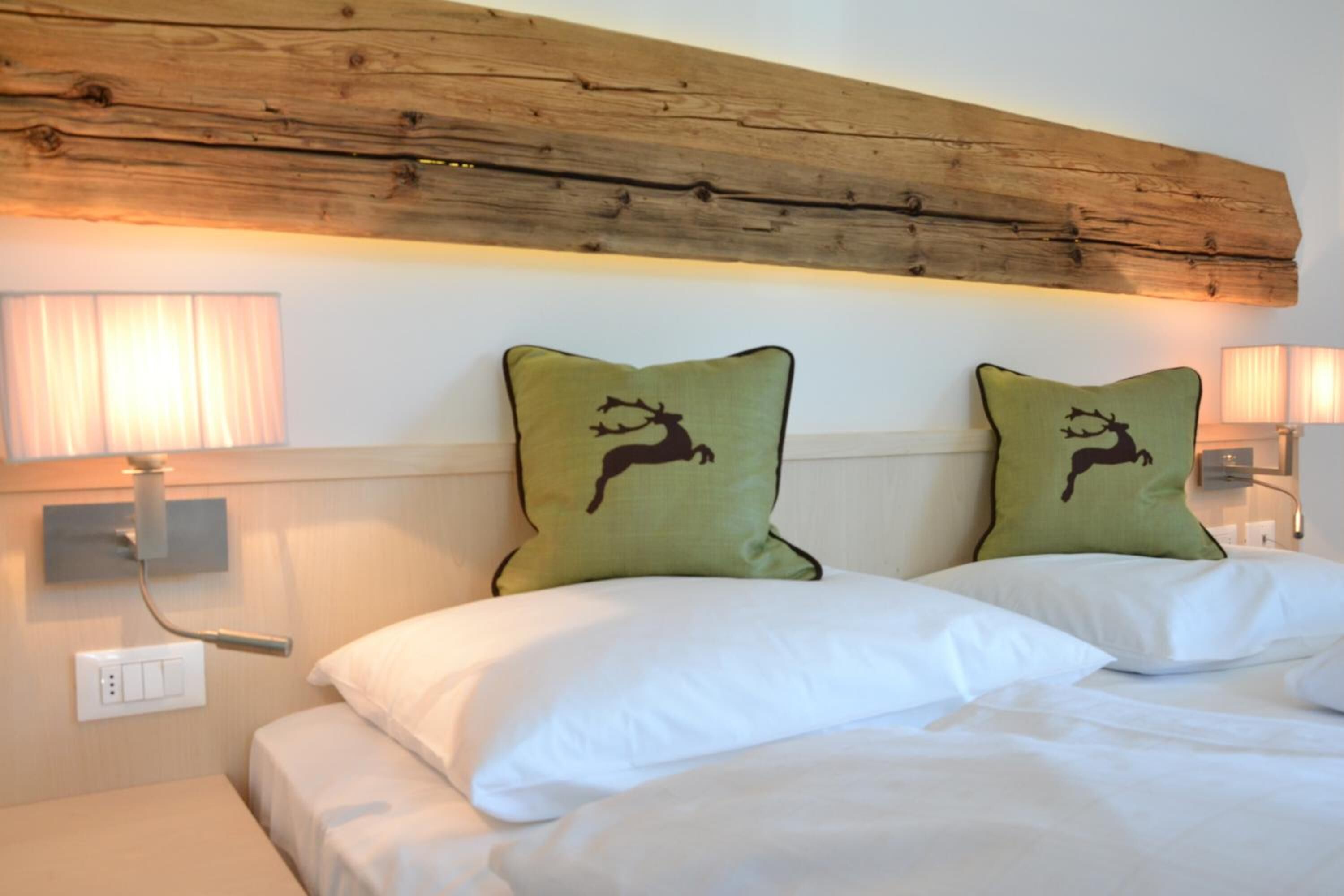 A hotel room with a wooden headboard, two green pillows with a deer design, white bedding, and bedside lamps.