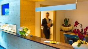 A hotel reception desk with a wooden panel wall, a computer monitor, and a vase with flowers. - Hotel Regglbergerhof (Nova Ponente)