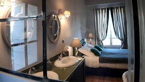 A hotel room with a large mirror, a bed with a headboard, a bedside table with a lamp, a window with curtains, and a chandelier.