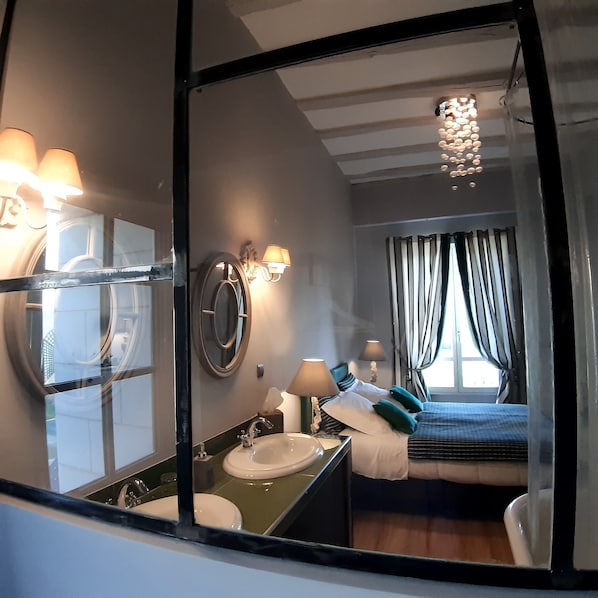 Habitación de hotel con un espejo grande, una cama con cabecero, una mesita de noche con lámpara, una ventana con cortinas y un candelabro.
