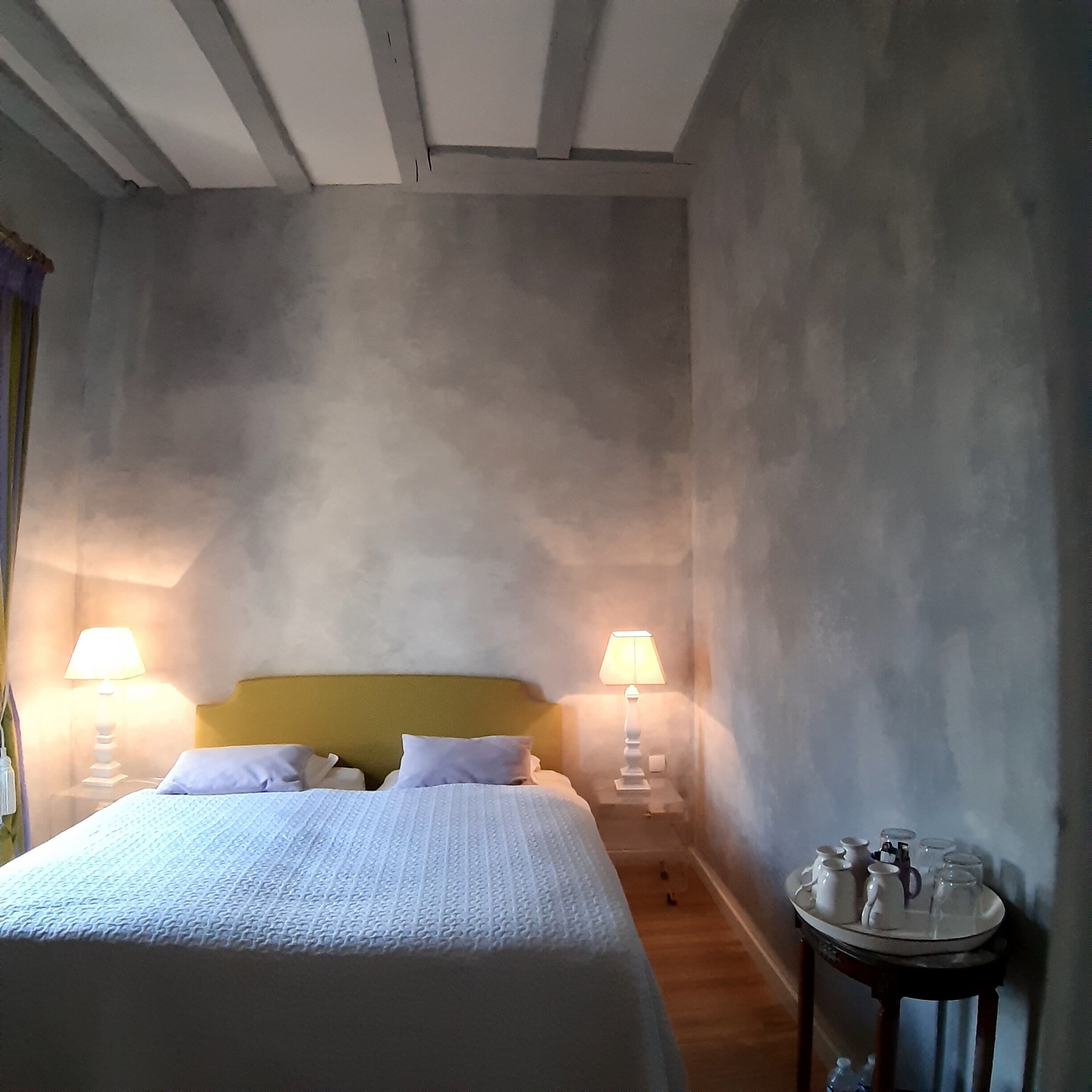 A bedroom with a bed, two bedside lamps, a nightstand with a tray of glasses, and a wooden floor.