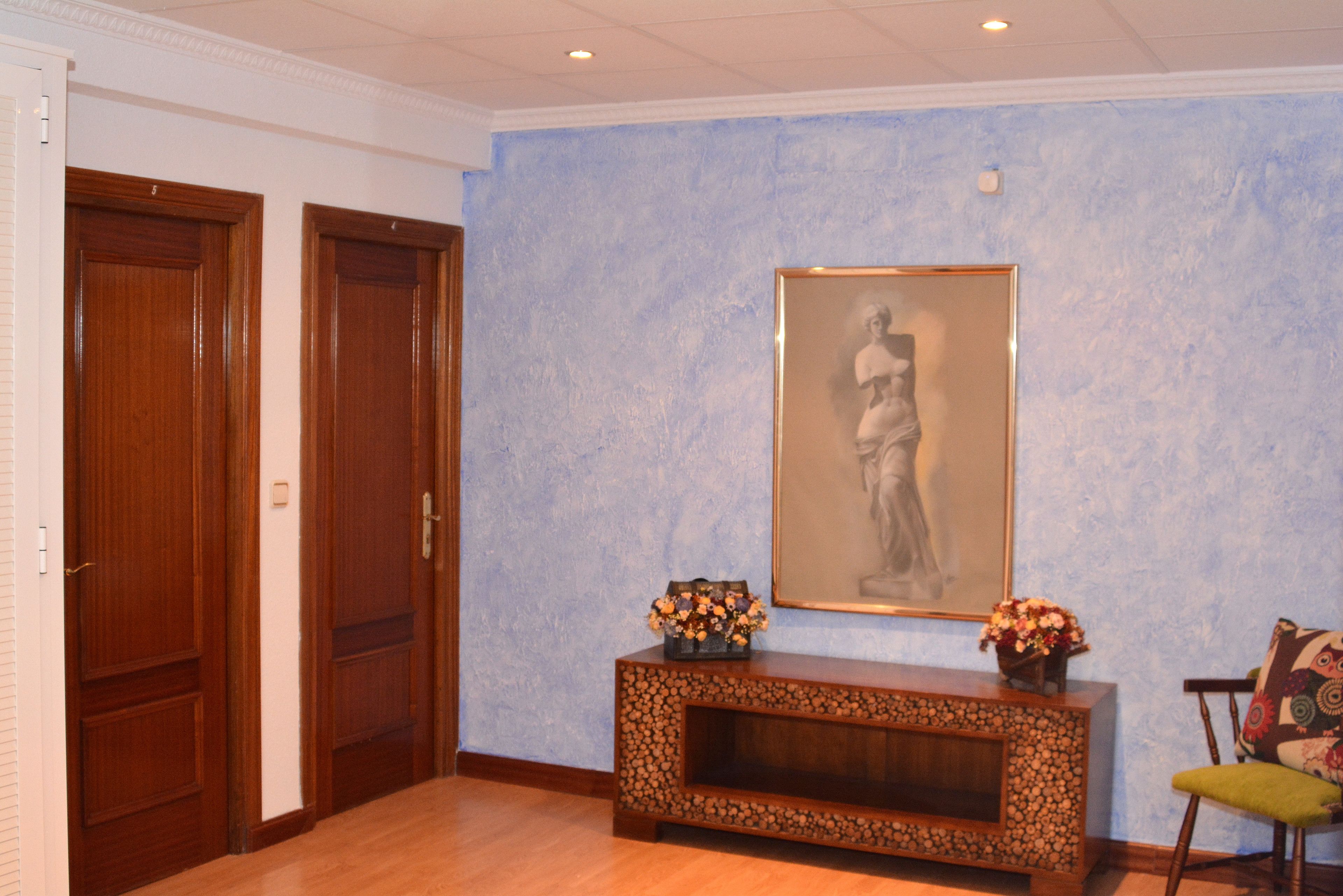 Ein Zimmer mit Holztüren, einer blauen Wand mit Struktur, einem gerahmten Kunstwerk, einem Konsolentisch mit Blumen und einem Sessel.