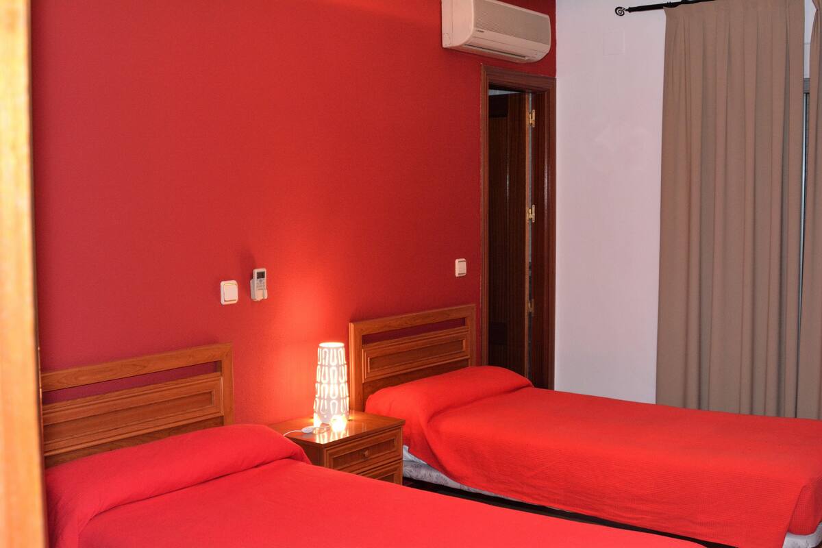 A hotel room with two beds, a nightstand with a lamp, an air conditioner, and a door leading to another room.