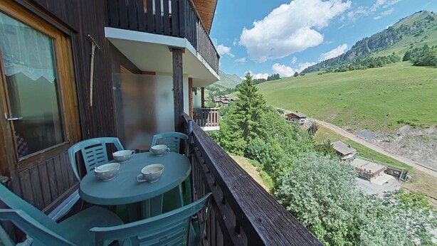 Un balcon avec une table et des chaises, donnant sur une vallée verdoyante et des montagnes lointaines.