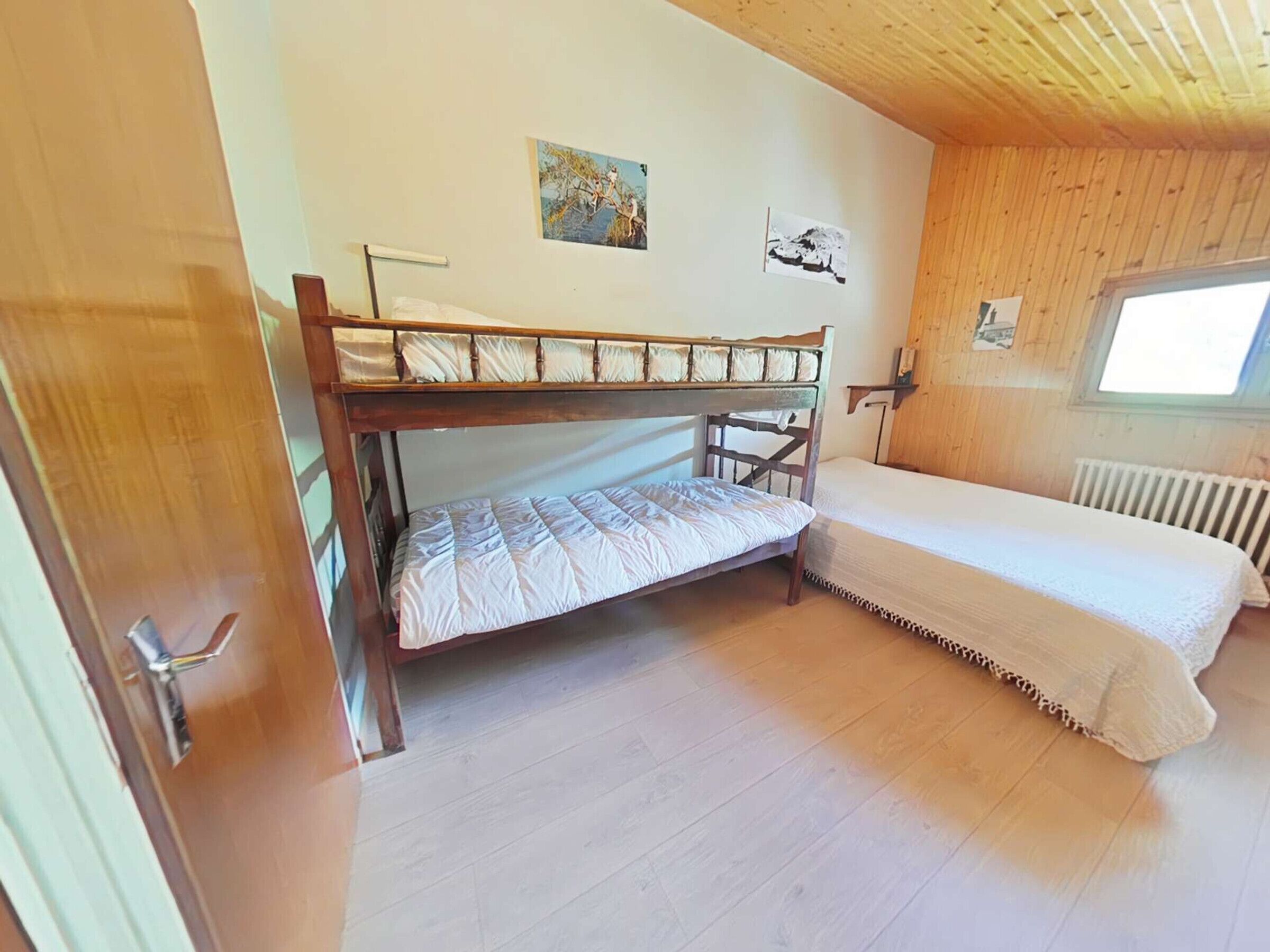 A room with a bunk bed, a single bed, wooden walls, a window, and a radiator.