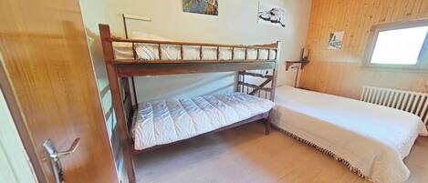 A room with a bunk bed, a single bed, wooden walls, a window, and a radiator.