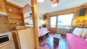 Une pièce compacte comprenant un coin cuisine, un espace repas avec une table et des chaises, un lit et une vue sur l’extérieur grâce à une grande fenêtre.