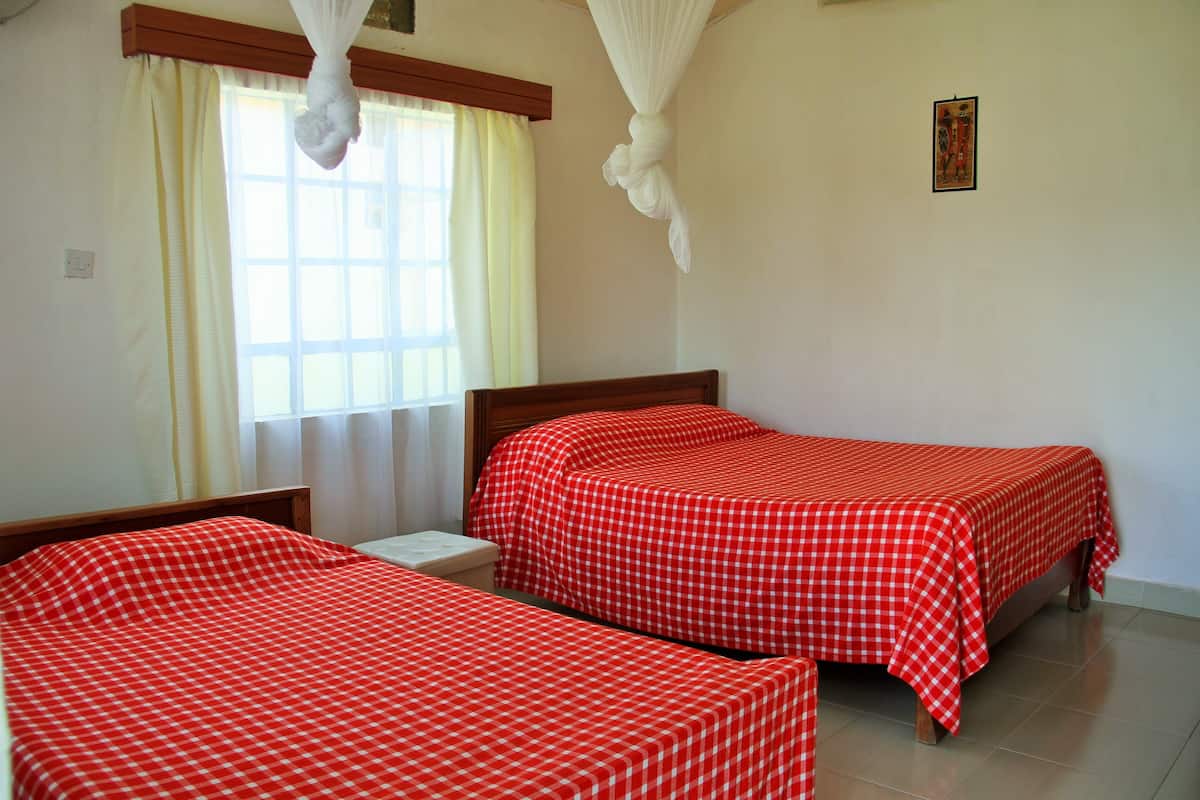 Quarto com duas camas, cada uma com uma colcha xadrez vermelha e branca. Há uma janela com cortinas, uma pequena mesa de cabeceira e um candeeiro fixado na parede.