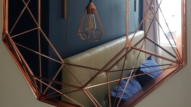 A geometric octagonal mirror with copper framing, reflecting a room with a blue wall, a hanging light fixture, and a white sofa with blue cushions. - Les Bruyères de Rouen - Seasonal rental (Sotteville-lès-Rouen)